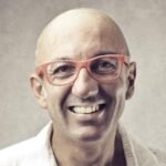 A bald, middle-aged man is smiling warmly at the camera. He is wearing large, red-framed glasses and a white shirt with the collar open. The background is a neutral gray, slightly textured, which contrasts with his bright expression and colorful glasses; perfect for a Post Page Template.