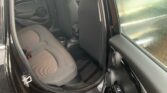 Interior of a black MINI viewed from the rear driver's side door. The back seats are gray with a fabric material. The door is open, revealing the wet glass window, likely from rain. The door panel has a cup holder, and the floor features a ribbed rubber mat.