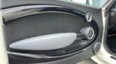 A close-up of a MINI car door interior, featuring a black curvy design element with a silver handle. The door has speakers integrated near the bottom and side. The background includes a blurred view through car windows, revealing part of an exterior building and pavement.
