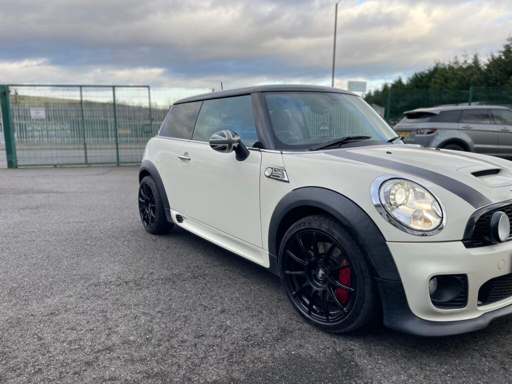A white MINI Cooper with black racing stripes on the hood is parked on an asphalt lot. The car sports black rims and red brake calipers. A metal fence and another gray vehicle are visible in the background under a cloudy sky.