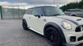 A white MINI Cooper with black racing stripes on the hood is parked on an asphalt lot. The car sports black rims and red brake calipers. A metal fence and another gray vehicle are visible in the background under a cloudy sky.