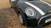 Close-up of a black MINI Cooper with white racing stripes parked on a gravel driveway. The front driver's side, including the headlight, grille, and part of the hood, is visible. The background shows part of a house and another car parked further away on the driveway.