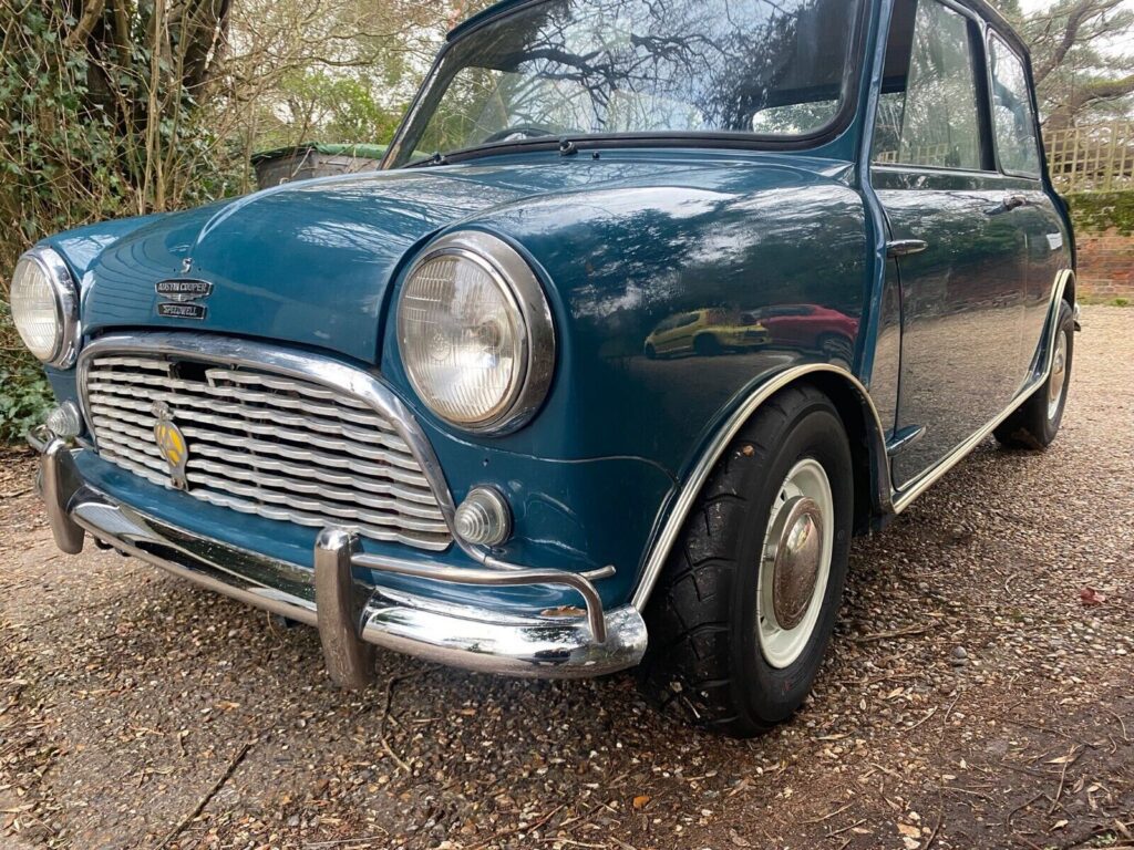 A classic dark blue MINI Cooper is parked on a gravel surface outdoors. The car has a rounded design with chrome trim, a distinctive front grille, circular headlights, and white sidewall tires. Trees and foliage are visible in the background, reflecting off the car's glossy surface.