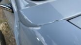 Close-up of a gray MINI's front end, focusing on the gap between the hood and the fender. The car's glossy surface reflects the sky. Part of the windshield and side mirror are visible. The hood and fender do not align perfectly, with a slight crack visible where the two panels meet.