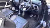 The interior of a MINI Cooper features a black and silver color scheme. It includes leather seats, a multifunctional steering wheel, a dashboard with round gauges, a center console with various controls, metal pedals, and door speakers. The driver's seat is on the right.