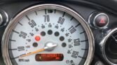 Close-up of a MINI car speedometer showing a top speed of 150 mph and a current reading at 0 mph. The odometer below the speedometer reads 43077 miles. The dashboard includes a triangular hazard light button on the right with a red triangle symbol and additional dashboard controls around.