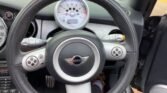 The interior of a MINI Cooper, focusing on the steering wheel. The wheel features an emblem in the center with the "MINI" logo. Behind it are the speedometer and tachometer. Visible controls adorn the steering wheel, while part of the car's door is open to the right.