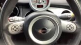 Close-up view of a MINI Cooper's steering wheel and dashboard. The steering wheel features the MINI logo and various control buttons. Behind the steering wheel, a central speedometer displays a reading of 165 mph, along with additional gauges and indicator lights.