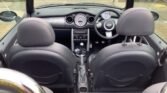 The interior of a MINI convertible car from a rear seat perspective. The car features black leather seats, a sleek dashboard with a central speedometer, and a three-spoke steering wheel. The gear shift is in manual mode, and the roof is retracted, offering an open view of the surroundings.