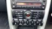 Close-up of a MINI's center console featuring a digital radio station display, climate controls with a central switch indicating 20°C, and various buttons and toggle switches. The radio displays "FM1 2, BBC, K2, TP." The console is a mix of black and metallic finishes.