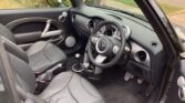 This image shows the interior of a MINI convertible with leather seats. The dashboard, steering wheel, gear shift, and pedal area are visible. The seats are black, and the dashboard features various dials and buttons. The car is parked on a street, with trees and a sidewalk visible outside.