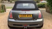 A metallic gray MINI Cooper convertible with a black soft top is parked in a driveway. The rear license plate reads "GU05 AYX". The driveway is paved with bricks, and there are trimmed hedges, a lawn, and a road visible in the background.