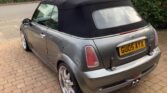 A gray convertible MINI Cooper with a black soft top is parked on a brick driveway. The vehicle has shiny alloy wheels and a visible exhaust pipe at the rear. The license plate reads "GU05 AYX." Nearby, a few cars and a hedge are seen along the street.