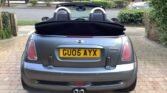 The image shows the rear view of a grey MINI convertible with a black soft top, parked on a brick driveway. The license plate reads "GU05 AYX." In the background are green shrubs, a tree, a stone wall on the left, and a residential street with additional greenery and house facades.