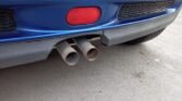 A close-up view of the rear bumper of a blue 2006 MINI Cooper S R53, showing dual exhaust pipes extending outward. The bumper includes a red reflector light and a black gridded vent. The car is parked on a concrete surface, showcasing part of the new clutch and its black underbody.