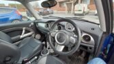 This image shows the interior of a 2006 R53 MINI Cooper S, featuring a black leather steering wheel with the manufacturer's emblem in the center. The dashboard includes several gauges and controls. The car seats have black upholstery. Outside, other vehicles and a building are visible. MOT till July.