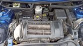 Close-up of an engine bay of a 2006 R53 MINI Cooper S. The engine components include various labeled parts such as the air filter box, coolant reservoir, and oil cap. The engine cover is prominently labeled "MINI" with various hoses and wiring visible, all set against a backdrop of a metallic blue car body. MOT till July.