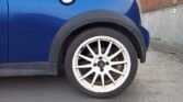 Close-up of a 2006 R53 MINI Cooper S rear wheel with a Team Dynamics Motorsport rim, showcasing a prominently white metal alloy wheel with multiple spokes and a black central hub. The car's wheel arch is visible, partially covered by black plastic fender trim. Background includes grey textured ground and part of a brick wall.