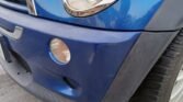 Close-up of the front right corner of a blue 2006 R53 MINI Cooper S, showcasing part of the headlight and bumper. The bumper has a round, clear fog light and a side marker. The car's tire and part of the fender are visible. The surface has minor scuffs, with slightly dull paint; clutch changed recently, MOT till July.