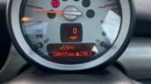 A close-up of a 2006 Mini Cooper S dashboard displays the speedometer and other indicators. The speedometer reads 0 mph, the car has driven 104,617 miles, and the temperature is 5.5°C. The tachometer shows RPM up to 8, with warning lights for the handbrake and another alert illuminated. Trees and grass are visible outside.
