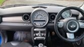 Interior view of a 2006 Mini Cooper with a black steering wheel featuring the Mini logo. The dashboard includes a large central speedometer, various control buttons, and two air vents. The manual gear shift is visible alongside cup holders in the center console. The driver's seat is black, complemented by a blue pan roof.