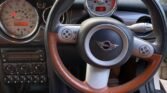 Image showing the interior of a 2008 Mini Cooper from the driver's perspective. The steering wheel has a chrome and brown leather design with the Mini emblem in the middle. The dashboard features a large central speedometer with smaller gauges. The seats are brown leather, ready to hit the sidewalk.