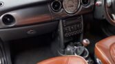 Interior view of a 2008 Mini Cooper 2dr featuring brown leather seats with vertical stitching. The dashboard of the 1.6 Cooper Sidewalk has a central speedometer, round air vents, and various control buttons. An air freshener with a smiley face hangs from the rearview mirror. The steering wheel and gear shifter are also visible.