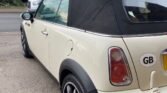 A cream-colored 2008 Mini Cooper convertible is parked on a street, with its black soft top roof closed. The car has a “GB” sticker on the rear, indicating Great Britain. Bare trees and another parked car are visible in the background. The ground is covered in gravel near the sidewalk.