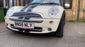 A white 2008 Mini Cooper with black racing stripes on the hood and license plate number BN08 NLY is parked in a driveway in front of a beige brick house with brown-framed windows. A green trash bin is visible on the left side near the house, just off the sidewalk. The car's front wheels are slightly turned to the right.