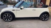 A white 2008 Mini Cooper 1.6 convertible is parked on the side of a residential street. It has a black soft-top roof, currently down, and stylish black alloy wheels. The interior features brown leather seats with headrests. A row of terraced houses is visible in the background.