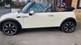 A white 2008 Mini Cooper convertible with a black retractable roof is parked on a driveway. The 1.6 Cooper features black and silver rims and brown leather seats, positioned near a brick house with the sidewalk in the background.