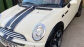 A white convertible 2008 Mini Cooper with black racing stripes is parked in a driveway. The car, featuring a black soft-top roof which is down and sleek black alloy wheels, could be the stylish Mini Cooper Sidewalk edition. A nearby green garbage bin and part of a brick building with windows and a door are visible in the background.