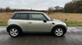 A silver 2008 Mini Cooper with black trim and a black roof is parked on a wet road. The car has silver alloy wheels and a visible antenna, accentuating its classic design. The background includes a grassy field, leafless trees, and an overcast sky, suggesting a dreary, wet day.