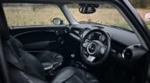 The image shows the interior of a 2008 Mini Cooper, highlighting its black leather seats, steering wheel, and dashboard. The view from the driver's seat provides a peek at the overcast outdoors through the wet window. The car's petrol-powered compact design emphasizes sleek lines and a minimalist console layout.