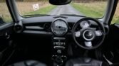 Interior view of a 2008 Mini Cooper’s cockpit, showcasing a black leather steering wheel, a central speedometer, and a smaller tachometer to the right. The dashboard features circular air vents, control dials, and a gear stick. Outside, a narrow rural road and grassy fields are visible.