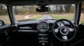 Interior view of a 2008 Mini Cooper, showcasing the driver's cockpit. The dashboard features a circular speedometer at the center, with tachometer and fuel gauge on either side. The steering wheel with the Mini logo is front and center. Beyond the windshield, there's a rainy landscape and a road sign. Experience this in the 1.6 Petrol variant.