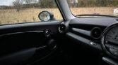 Interior of a 2008 Mini Cooper, showing the dashboard and front passenger seat area. The dashboard features round air vents and a large circular display. The side mirror is visible through the rain-speckled window. Outside the car, a grassy field with scattered trees extends into the background.