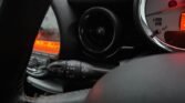 Close-up view of a 2008 Mini Cooper dashboard with a focus on the turn signal and light control lever. The red backlighting reveals the tachometer and other controls, while the air vent is also visible near the lever. The image conveys a dark and sleek interior typical of a Mini Cooper 1.6.