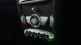 Close-up of a 2008 Mini Cooper's control panel illuminated by green and red lights. The central dial is surrounded by various symbols and smaller buttons. Below the central dial, five toggle switches with green indicator lights manage functions efficiently. The panel is bordered by a dark dashboard, embodying classic petrol-driven charm.