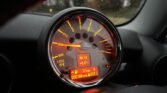 Close-up of a 2008 Mini Cooper dashboard illuminated in red and orange. The speedometer's needle points just above 1 (x1000 RPM), with various icons visible. An embedded digital display shows the time (16:12), temperature (5.0°C), trip distance (37 miles), and total mileage (90,198 miles) for this petrol-fueled car.