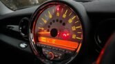 The image shows a close-up view of a 2008 Mini Cooper's speedometer and dashboard, illuminated with orange-red lighting. The speedometer displays large numbers ranging from 0 to 160. Below the speedometer are controls for the radio/CD player, AUX input, and several circular buttons and knobs.