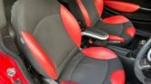 The interior of a MINI car showcases two front seats upholstered in black and red fabric and leather. The car door, partially visible, is also black and red, matching the seats. The armrest between the seats is black with a silver top, and the steering wheel is partially visible on the right.