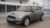 A gray MINI Cooper with a UK license plate BV59 OHG is parked indoors in a bright, modern space. The car has silver alloy wheels, a black roof, and a hood scoop. The background features large windows, railings, and sleek architectural elements.