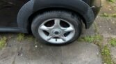 A black MINI car with a flat tire is parked on a stone surface, surrounded by patches of grass and dirt. The tire rim prominently displays the "MINI" logo. Evidence of recent rain is apparent through the damp ground, while the tire and fender appear slightly dirty.