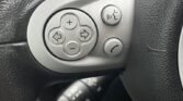 Close-up of a MINI steering wheel equipped with control buttons. On the left side, buttons include volume up, volume down, and navigation arrows in a circular arrangement. Below these, there are two additional buttons for hands-free phone and voice commands. The speedometer is partially visible in the background.