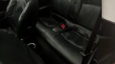 Interior view of a MINI's back seat area. The seats are black leather with red seat belt accents. The front passenger seat is partially angled towards the rear. There's a visible seatbelt buckle on the rear seat. The car door is open, showing the door jamb and a ground cover of pebbles.