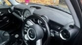 The image shows the interior of a black MINI with leather seats, displaying the steering wheel, dashboard, and front seats. A pink smiley face air freshener hangs from the rearview mirror. Outside the car, a brick wall and a window of a building are visible. The left door is slightly open.