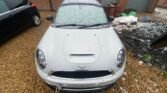 A white MINI COOPER 2.0 SD Coupe with a tow hook is parked on a gravel surface next to a black car. The windshield and roof are partially covered with snow. In the background, there are brick buildings with metal roller doors, and some construction materials, including white bags and green tarps, are scattered around.