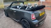 A gray MINI convertible parked on a gravel surface, with its roof down. The car has a UK license plate reading "AK61 RXJ." The interior features black and white seats, and the car boasts sporty alloy wheels. A brick building and trees are visible in the background.