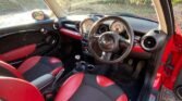 The interior of a red MINI compact car features black and red leather seats, a black steering wheel, and a dashboard with round gauges. The dashboard includes air vents, a central display, and control knobs. Sunlight streams through the windows, illuminating the clean, modern design.